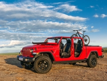Jeep Gladiator na cor vermelha em terreno arenoso, sem portas e teto, com para-brisa rebatido e bicicleta na caçamba. O solo é arenoso e ao fundo um céu azul claro com nuvens.