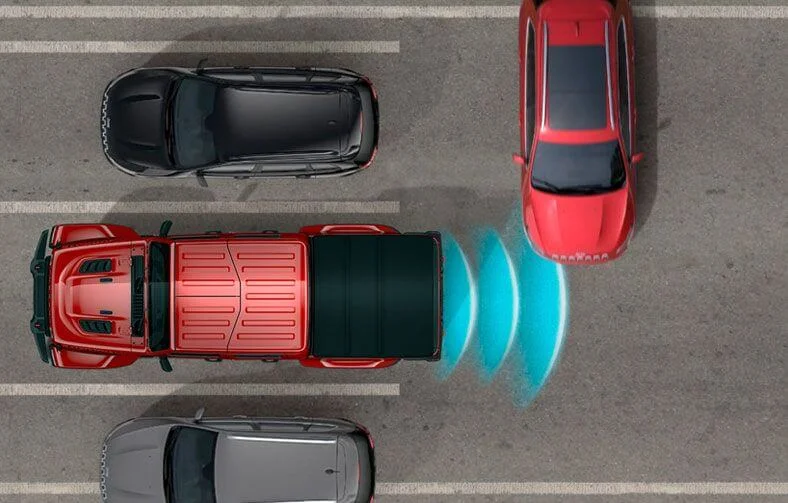 Linhas circulares na traseira do Jeep Gladiator visto de cima simulam o detector de tráfego cruzado durante a saída de estacionamento entre dois veículos na horizontal, com um carro vermelho na vertical se aproximando da traseira.