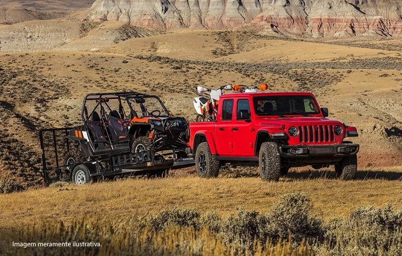 Jeep Gladiator com duas motos na caçamba e reboque de com quadriciclo em terreno com grama baixa.