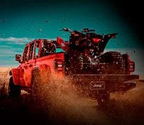 Picape Jeep Gladiator vermelha vista de trás, com quadriciclo na caçamba aberta, em terreno arenoso.