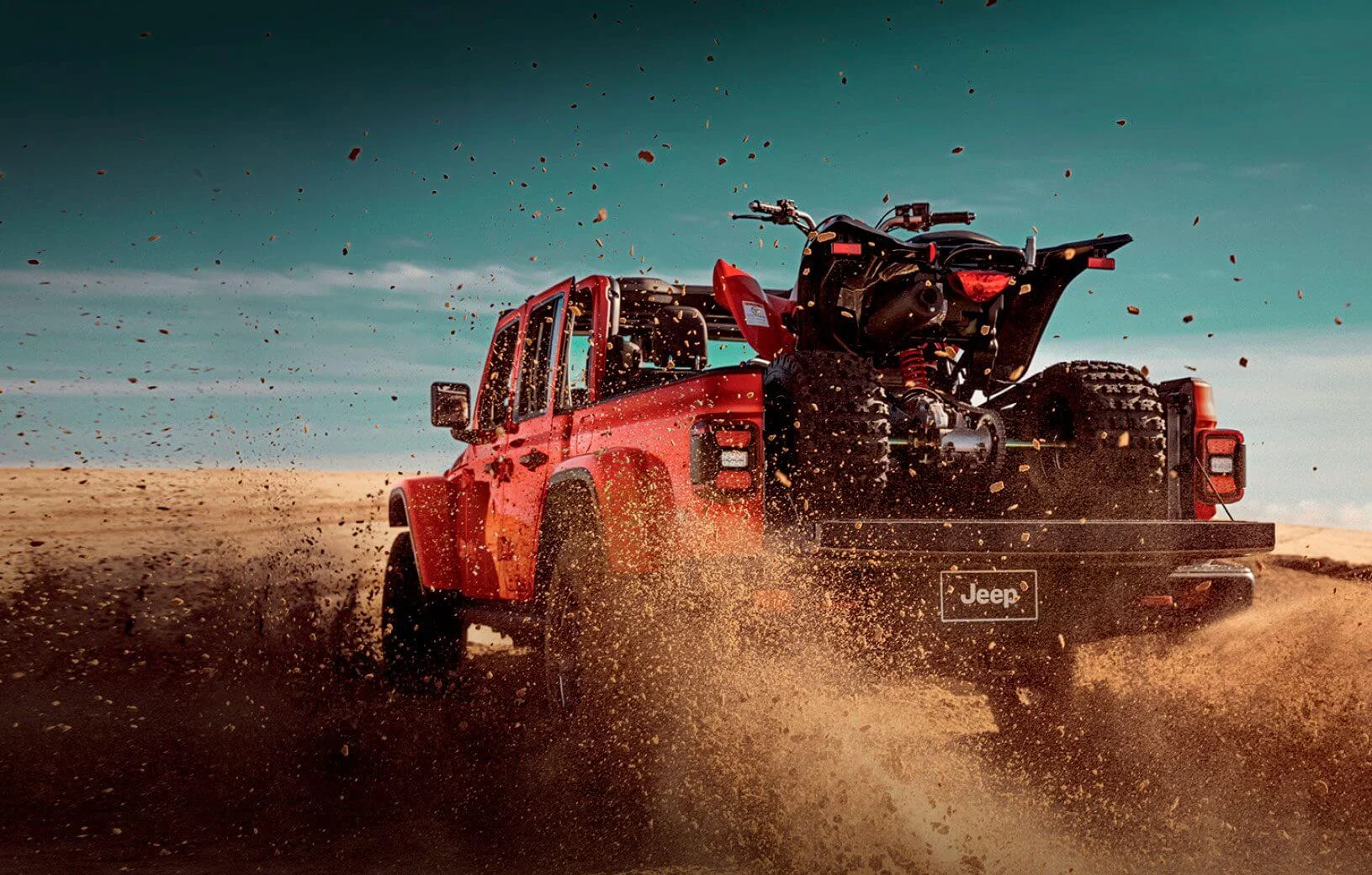 Picape Jeep Gladiator vermelha vista de trás, com quadriciclo na caçamba aberta, em terreno arenoso.