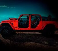 Picape Jeep Gladiator vermelha sem portas e teto, com o para-brisa rebatido em pista de terra e cascalho, com montanhas e céu claro sem nuvens ao fundo.