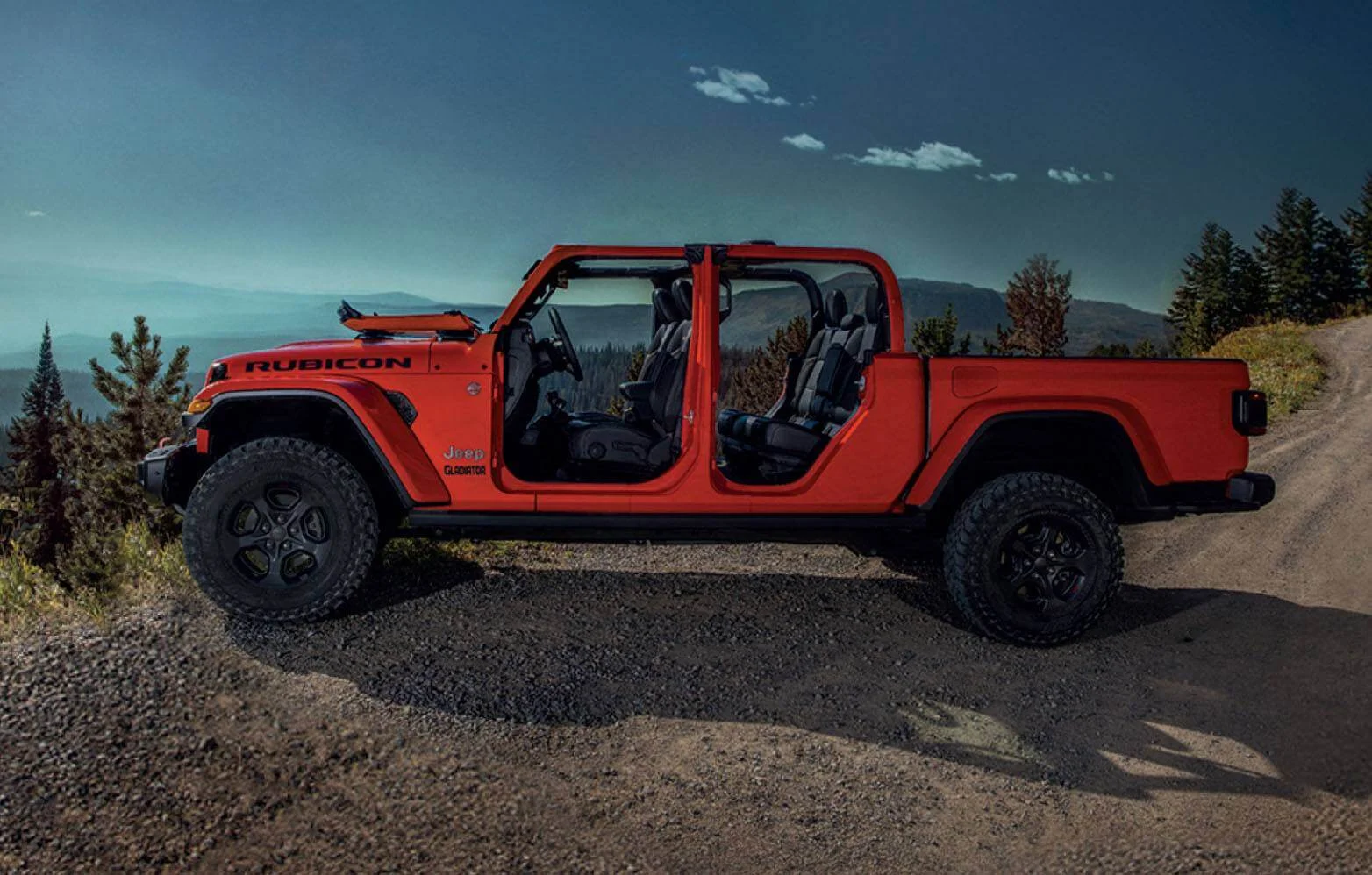 Picape Jeep Gladiator vermelha sem portas e teto, com o para-brisa rebatido em pista de terra e cascalho, com montanhas e céu claro sem nuvens ao fundo.