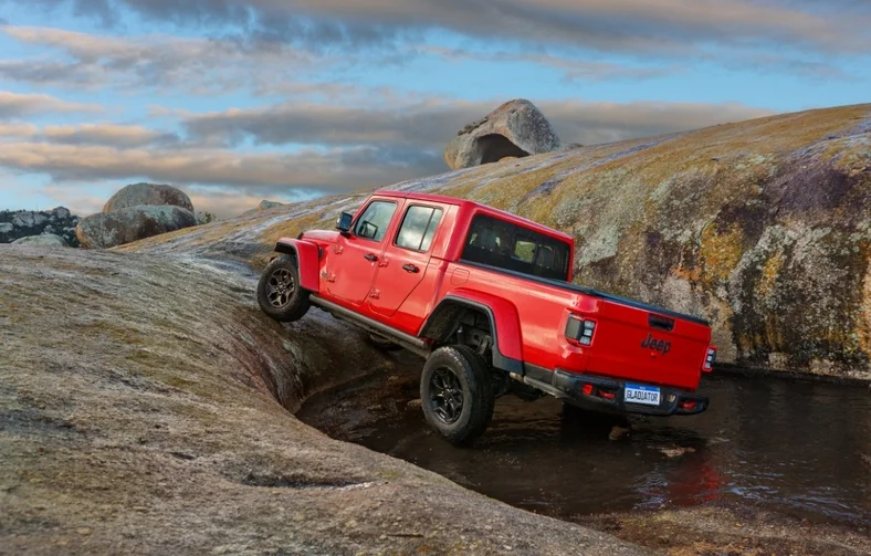 Picape Jeep Gladiator vermelha vista de trás saindo de lago e subindo em terreno rochoso.